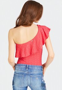 Haut rouge à une épaule avec manche à volants, motif floral texturé, associé à un jean bleu clair avec poches et broderie florale sur un côté.