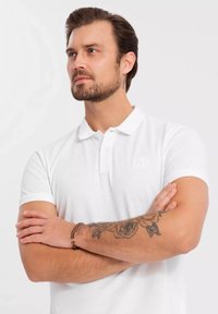 Bílá bavlněná polo triko s texturovaným povrchem, které má dvouknoflíkovou légu a diskrétní logo na hrudi. Model předvádí tetovanou ruku.