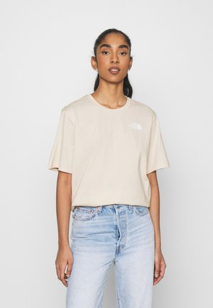 T-shirt di cotone beige a maniche corte, con scollo rotondo, caratterizzato da un piccolo logo bianco sul lato sinistro del petto, abbinato a jeans di denim blu chiaro.