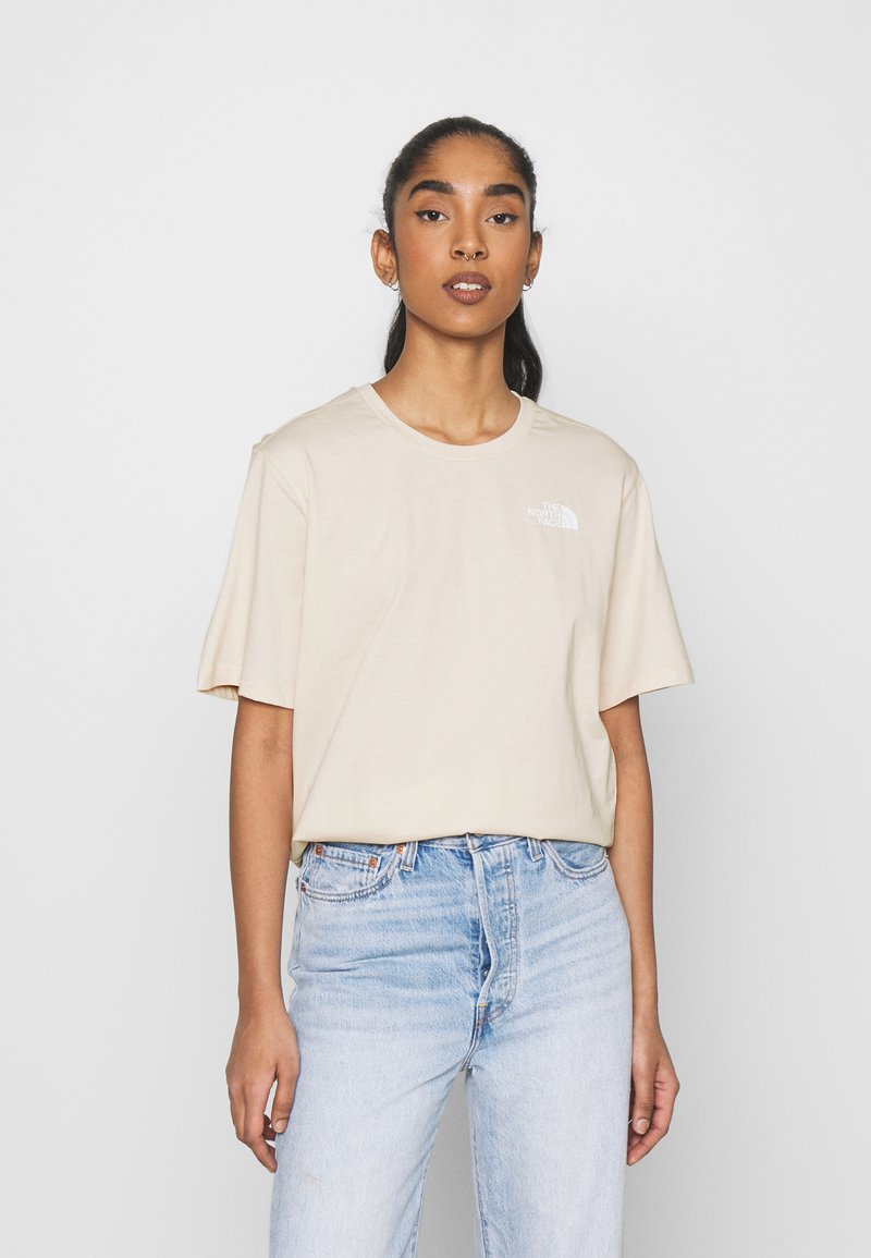 T-shirt di cotone beige a maniche corte, con scollo rotondo, caratterizzato da un piccolo logo bianco sul lato sinistro del petto, abbinato a jeans di denim blu chiaro.
