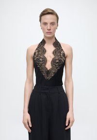 Άτομο που φοράει μαύρο αμάνικο τοπ με βαθύ δαντελένιο V-λαιμόκοψη και μαύρο παντελόνι με ψηλή μέση, στέκεται μπροστά σε απλό φωτεινό φόντο.