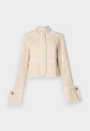 Beige zugeschnittene Jacke aus weichem Material, mit weißen Fransenakzenten entlang der Kanten und großen dekorativen Knöpfen an den Ärmelabschlüssen.