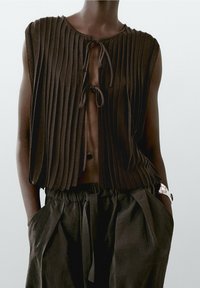 Brown pleated top with a tie closure at the neckline, paired with loose, high-waisted black pants. Textured fabric and casual design.