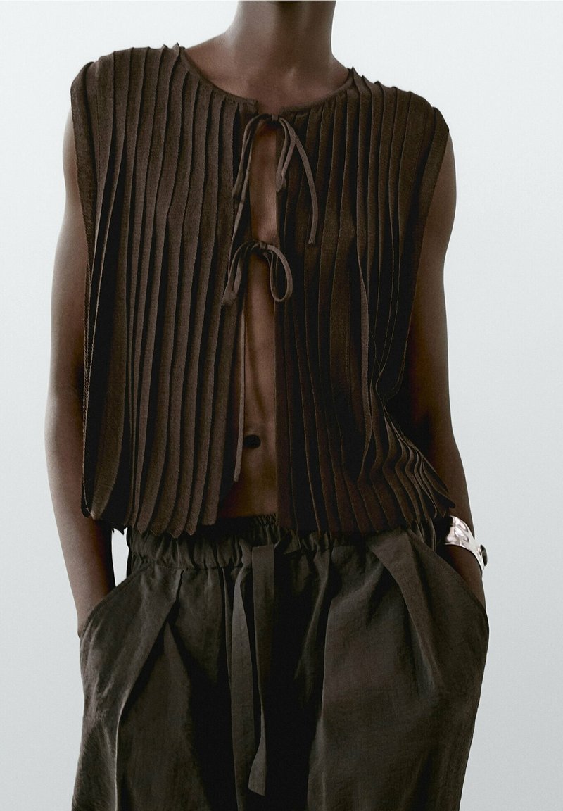 Brown pleated top with a tie closure at the neckline, paired with loose, high-waisted black pants. Textured fabric and casual design.