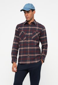 Geruite flanellen shirt in diep maroon en marineblauw, met lichtbruine accenten. Twee borstzakken en een knoopsluiting aan de voorkant. Draagt een lichte denim pet.