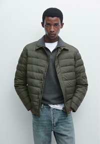 Olive green puffer jacket with a smooth texture, zip front, and raised collar, layered over a grey sweater and white t-shirt, paired with blue jeans.