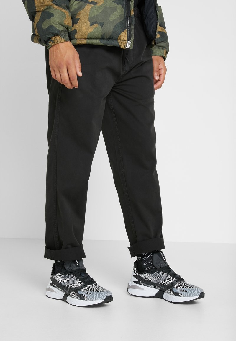 Pantaloni neri con risvolti, abbinati a sneakers nere e grigie con tomaia in rete e suola bianca. Giacca puffer camouflage visibile.