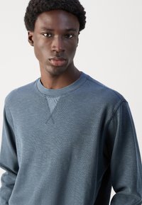 Ανδρικό μπλε βαμβακερό sweatshirt με στρογγυλή λαιμόκοψη, μακριά μανίκια, υφή υφάσματος και λεπτομέρεια σταυροβελονιάς στην ραφή του λαιμού.