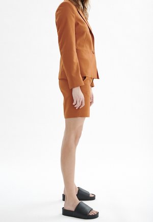 Oranžové přizpůsobené sako a odpovídající šortky, doplněné černými nazouvacími sandály. Hladká textura a přiléhavý střih, zobrazeno z boku.