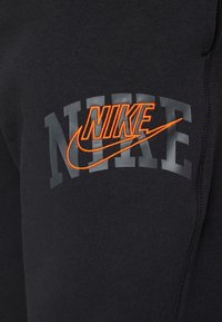 Schwarzer Stoff mit grauem "NIKE"-Text und einem überlappenden orangefarbenen, bestickten Nike-Logo und Swoosh.