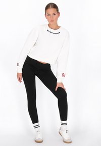 Sudadera blanca con texto bordado en negro, combinada con leggings negros ajustados y zapatos blancos con detalles beige y calcetines negros a rayas.