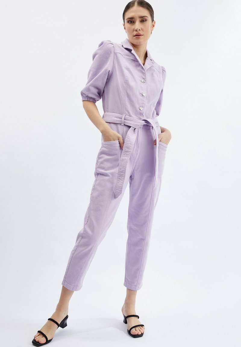 Lila jumpsuit met korte pofmouwen, knoops sluiting, riem in de taille en zijzakken. Draagt met zwarte open sandalen met hakken.
