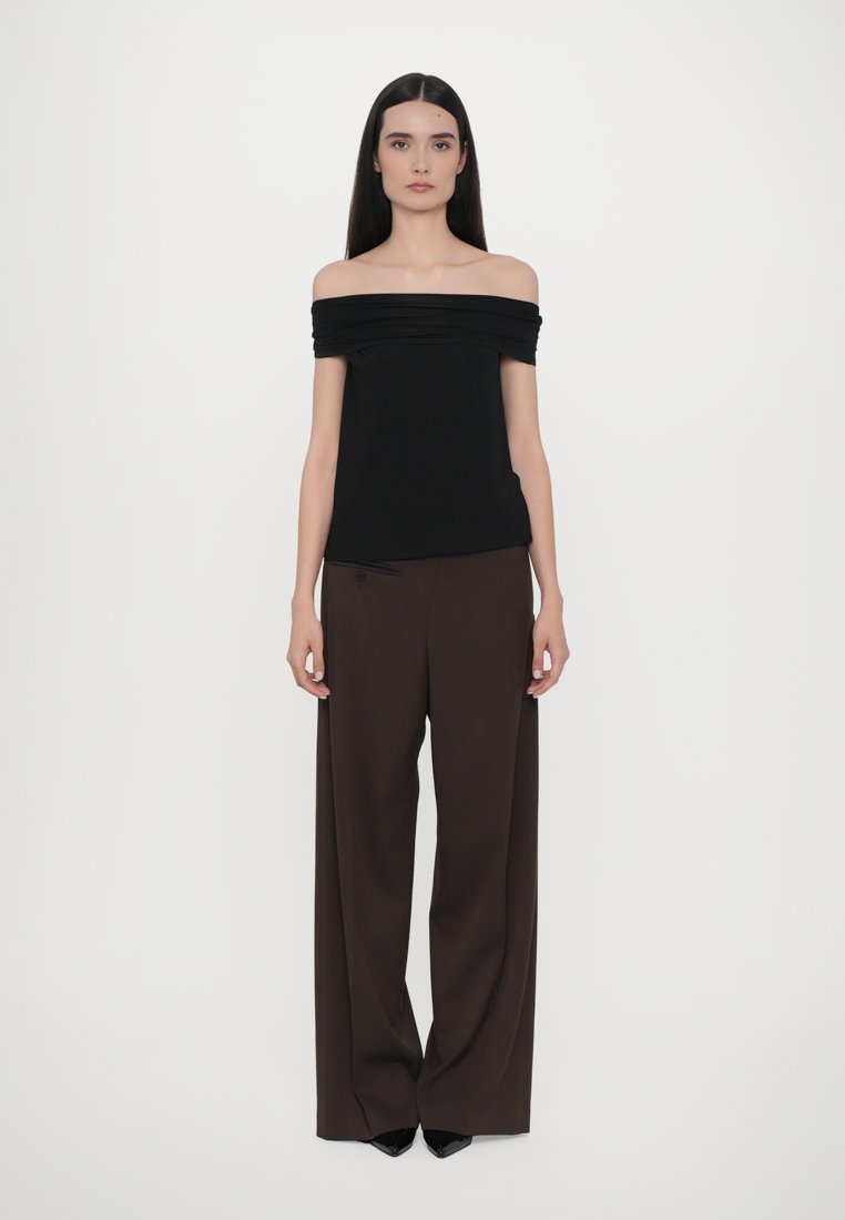 Femme aux longs cheveux noirs portant un haut noir à épaules dénudées et un pantalon marron à jambes larges, se tenant devant un fond blanc uni.