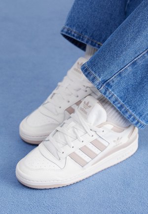 Hvide Adidas sneakers med beige striber, båret med hvide sokker og blå jeans på et blåt tæppe.