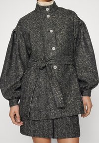 Textured, woven fabric jacket in dark colors with metallic fibers. Features a high collar, large buttons, and a tied belt at the waist.