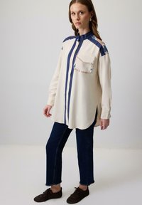 Crème langemouwenshirt met marineblauwe accenten en een voorzak, met een gebogen zoom en knoopsluiting, gecombineerd met donkerblauwe jeans.