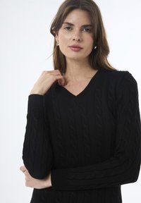 Giovane donna con capelli castano chiaro che indossa orecchini d'argento e un maglione a V con lavorazione a trecce nero, su uno sfondo bianco uniforme.