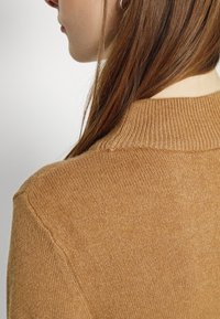 Brun stickad tröja med en ribbad krage och mjuk textur, visad från en närbild bakifrån med ett mjukt, lättviktigt tyg.