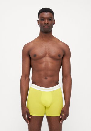 Calvin Klein gele boxershorts met een gestructureerde stof, aansluitende pasvorm en een witte logo tailleband, gepresenteerd op een model met een neutrale achtergrond.