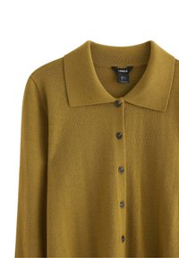 Sennepsfarvet cardigan lavet af struktureret strikstof. Har krave, knaplukning foran og mørke knapper. Etiketten er synlig inde i kraven.