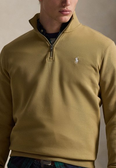 Vyras dėvi garstyčių geltonumo ketvirčio užtrauktuko megztinį su mažu baltu siuvinėtu logotipu kairėje krūtinės pusėje, dėvi tamsiai marškinius po juo.