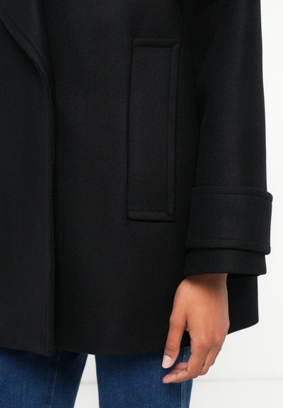 Close-up de uma pessoa a usar um casaco preto com um bolso lateral retangular e mangas com punho, combinado com jeans azuis.
