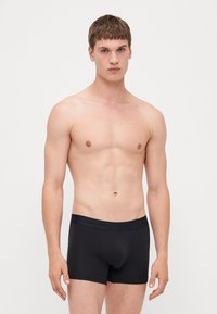 Schwarze Boxershorts aus Bio-Baumwolle mit glatter Textur und einem weichen Bund, der mit "Organic Basics" beschriftet ist. Das Modell ist ohne Oberteil zu sehen.