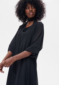 Kobieta o kręconych włosach, ubrana w czarną sukienkę z szerokimi rękawami i pasujący do niej choker, patrząca lekko w lewo na tle jednolitego tła.