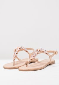 Rose gold flat sandals with a toe post design, featuring a decorative strap of clear and pink gemstones. Smooth, shiny material and minimal sole.