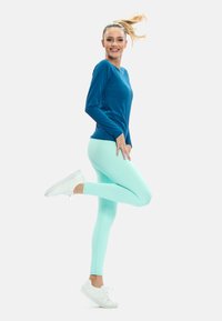 Långärmad blå idrottströja matchad med mintgröna leggings. Vita sneakers har en enkel design. Håret är uppsatt, kvinnan är i rörelse.