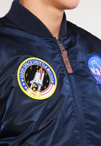 Tumesinine bomber-jakk, valmistatud satiinkangast, millel on kaks broderi embleemi: NASA logo ja kosmosesüstiku missiooni embleem.