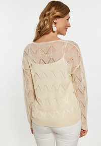 Pulover bej tricotat cu un model în zigzag, textură cu țesătură deschisă și decolteu larg. Asortat cu pantaloni albi.
