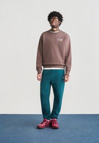 Καφέ φούτερ με κεντημένο λογότυπο, πράσινο μπλε φόρμας παντελόνι και ροζ αθλητικά παπούτσια. Το ύφασμα είναι μαλακό· το σχέδιο έχει χαλαρή εφαρμογή με ριμπ μανσέτες.