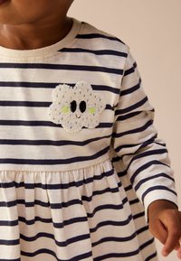 Abito a maniche lunghe a righe beige e blu navy con vita arricciata. Presenta una toppa a forma di nuvola soffice con occhi e dettagli verdi sul petto.