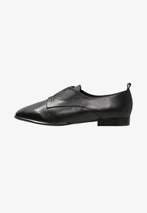 Zapato de cuero negro con punta afilada, diseño sin cordones, parte superior texturizada y tacón bajo y plano. Presenta costuras sutiles y una pequeña lengüeta.