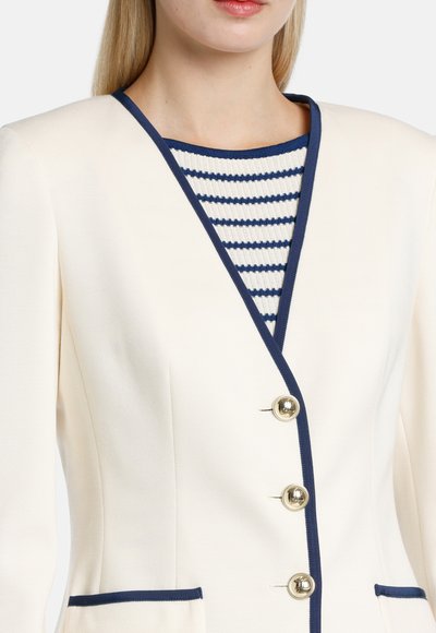 Ivory blazer dengan trim dan kancing navy, dilengkapi dengan saku depan. Di bawahnya, terdapat kemeja bergaris biru dan putih yang sebagian terlihat.