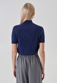 Donna con lunghi capelli biondi che indossa un maglione corto e peloso blu navy a maniche corte e pantaloni grigi a vita alta, di spalle su uno sfondo bianco.