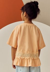 Pfirsichfarbendes Baumwoll-T-Shirt mit kurzen Ärmeln, Spitzenbesatz am Saum und einem Rüschensaum. Die Rückansicht zeigt eine lässige Passform und ein schlichtes Design.
