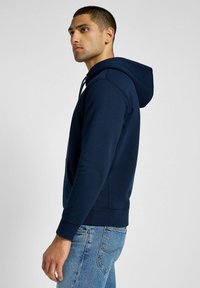 Granatowy hoodie z mieszanki bawełny, z regulowanym kapturem, przednią kieszenią typu kangur, ściągaczami na rękawach i luźnym krojem. Nosi się go z niebieskimi dżinsami.