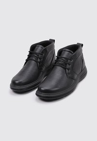 Un par de botas de cuero negro hasta el tobillo con delanteras con cordones y cuellos acolchados sobre un fondo gris.