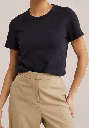 Vrouw draagt een effen zwart T-shirt met korte mouwen, in beige hooggetailleerde broek gestopt, poseert tegen een neutrale achtergrond.