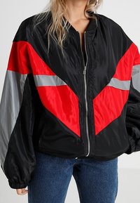 Giacca bomber nera con strisce a chevron rosse e grigie, materiale in nylon lucido, chiusura con zip, vestibilità oversize e polsini elasticizzati.