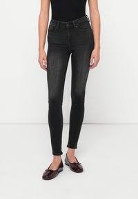 Jeans negri din denim cu talie înaltă, cu o formă ajustată. Prezintă buzunare și detalii ușor decolorate. Sunt asortați cu mocasini burgund.