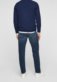 Homme portant un pull bleu foncé, un jean bleu, des baskets blanches et une chemise blanche, debout de dos sur un fond uni.