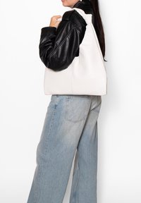 Borsa a tracolla in pelle bianca con forma arrotondata e tessitura morbida. Indossata con giacca in pelle nera e pantaloni di jeans azzurri.