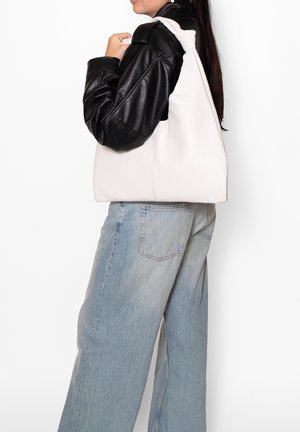 Borsa a tracolla in pelle bianca con forma arrotondata e tessitura morbida. Indossata con giacca in pelle nera e pantaloni di jeans azzurri.