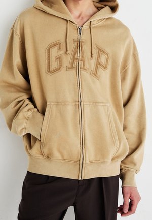 Beiger Kapuzenpullover mit Reißverschluss und Fronttaschen sowie „GAP“-Logo, getragen von einer Person mit einer Hand in der Tasche und der anderen entspannt an der Seite.