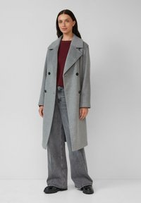 Vrouw in een lange grijze dubbelzijdige jas, bordeauxrode trui, grijze jeans met wijde pijpen en zwarte schoenen, staande tegen een effen muur.