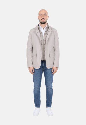 Uomo con testa rasata e barba in piedi, indossa giacca beige sopra gilet grigio con zip, camicia bianca, jeans blu e scarpe da ginnastica bianche.