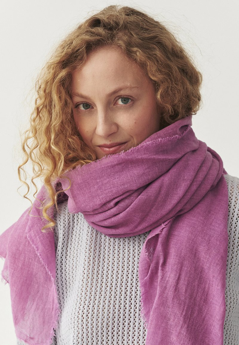 Écharpe en lin violet enroulée autour du cou, avec un tissage texturé et des bords effilochés, complétant un haut en maille ouverte gris clair.